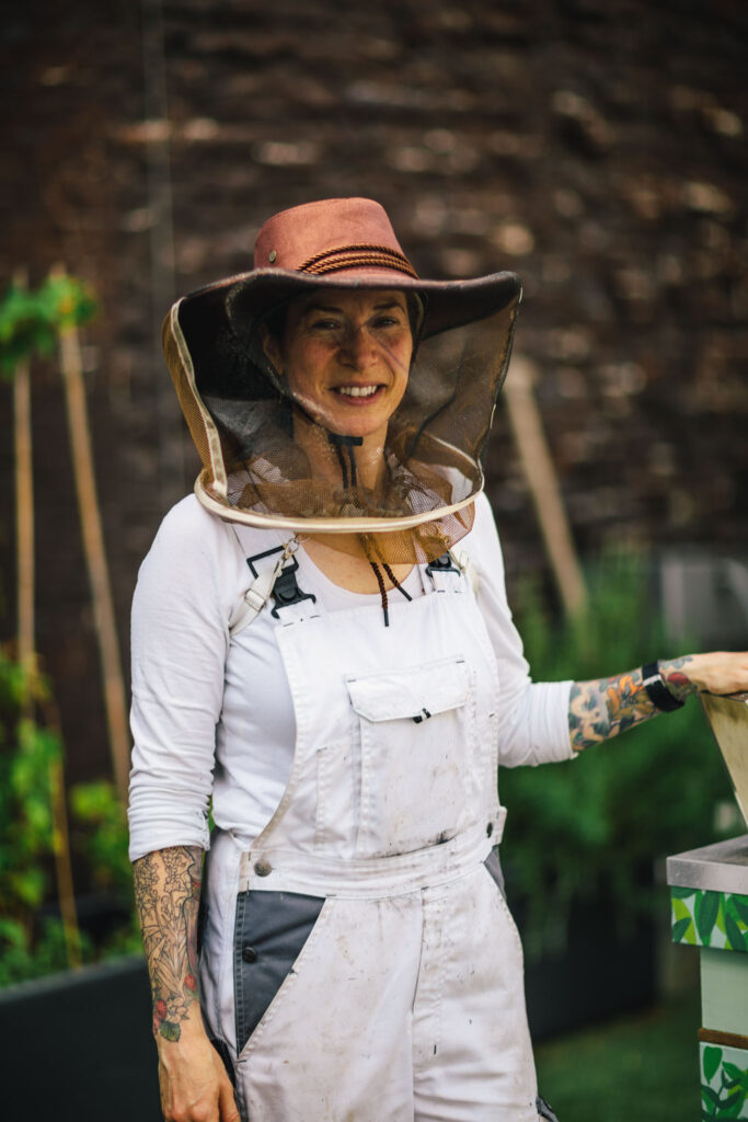 Anoushka de Graaf, beekeeper at The Dylan Amsterdam, posing proudly next to her beehive, dressed in an overall and with an beekeeper's hat.